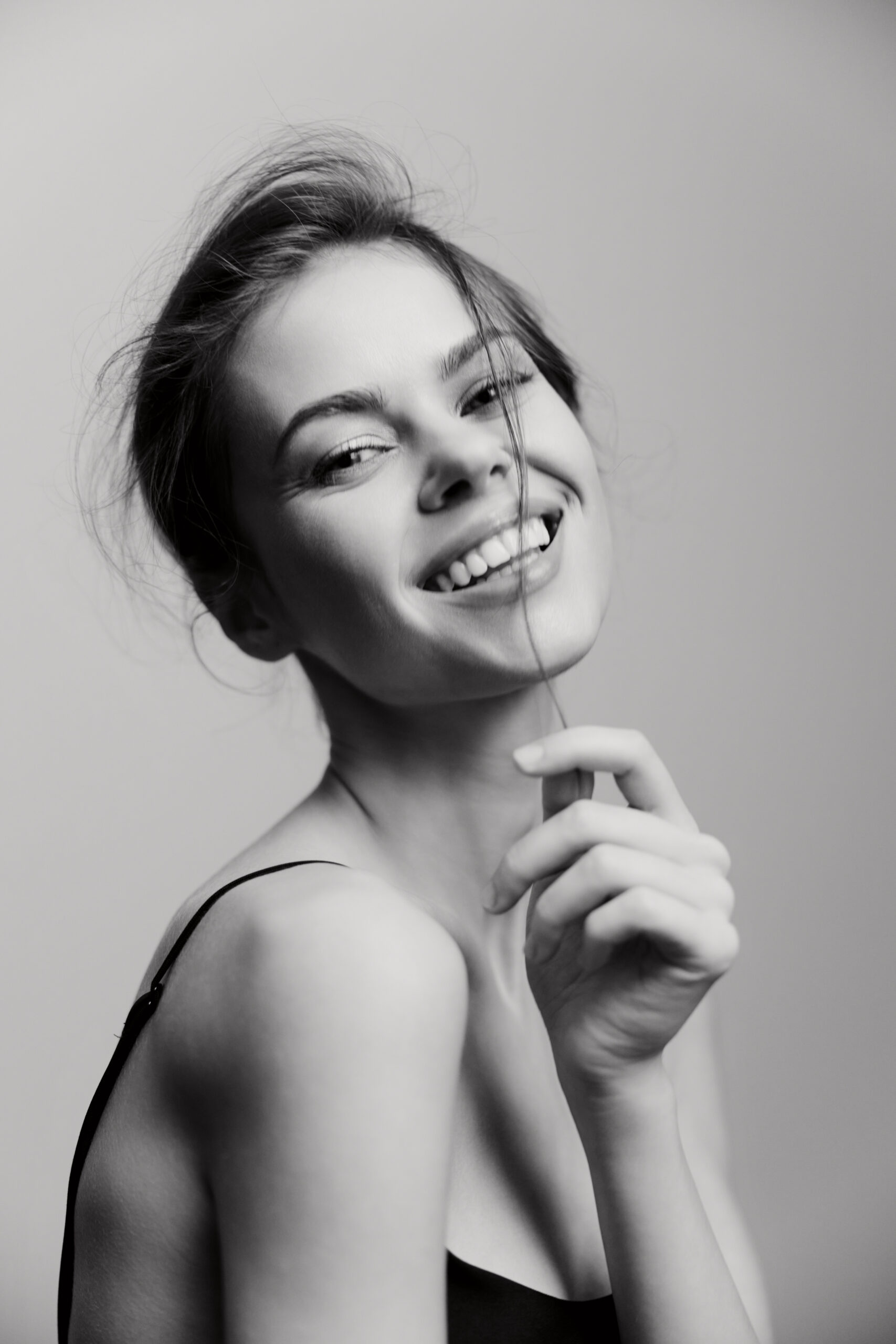 Joyful young woman smiling brightly, playing with her hair, captured in a black and white portrait against a minimalistic background. Emotions and beauty concept.